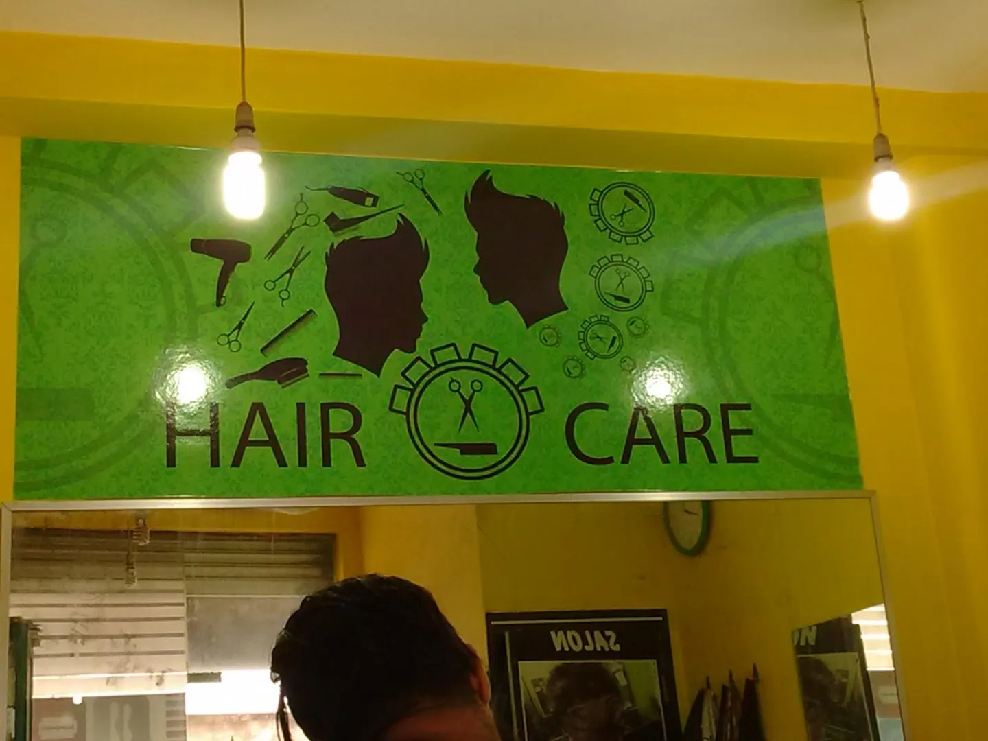 Hair Care interior signage panel: lime-green background with two black male profile silhouettes, scissors, comb, razor, and dryer icons, and the HAIR CARE wordmark with a scissor-and-comb emblem